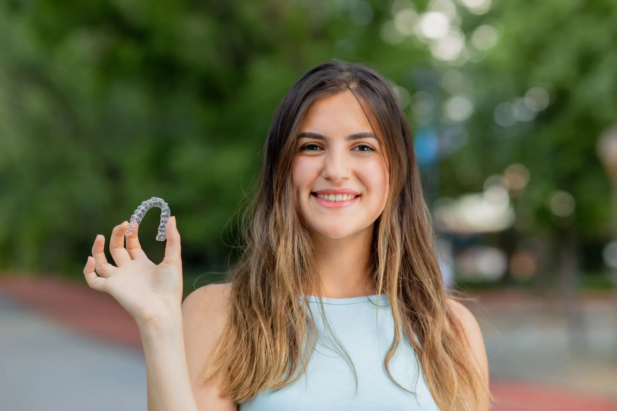 Smiling teenage girl holding Invisalign aligner outdoors, showcasing comfortable orthodontic treatment option for teens in St. Augustine, FL.
